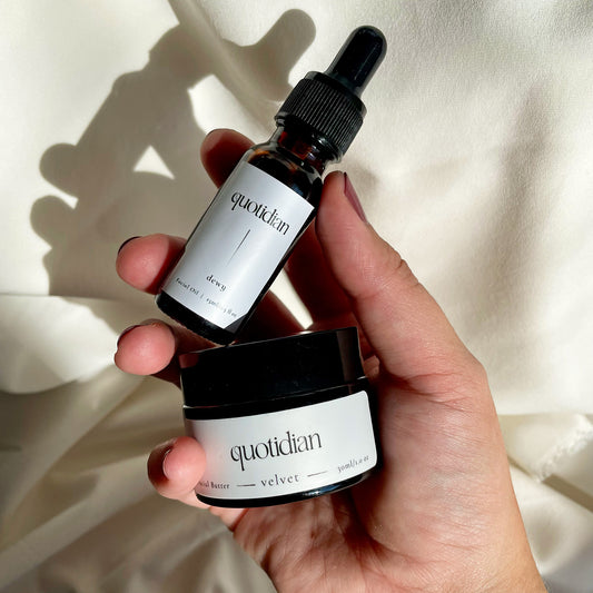 The image shows a hand holding a bottle of Quotidian Dewy Face Oil and a jar of Quotidian Velvet Face Butter against a soft, neutral background.